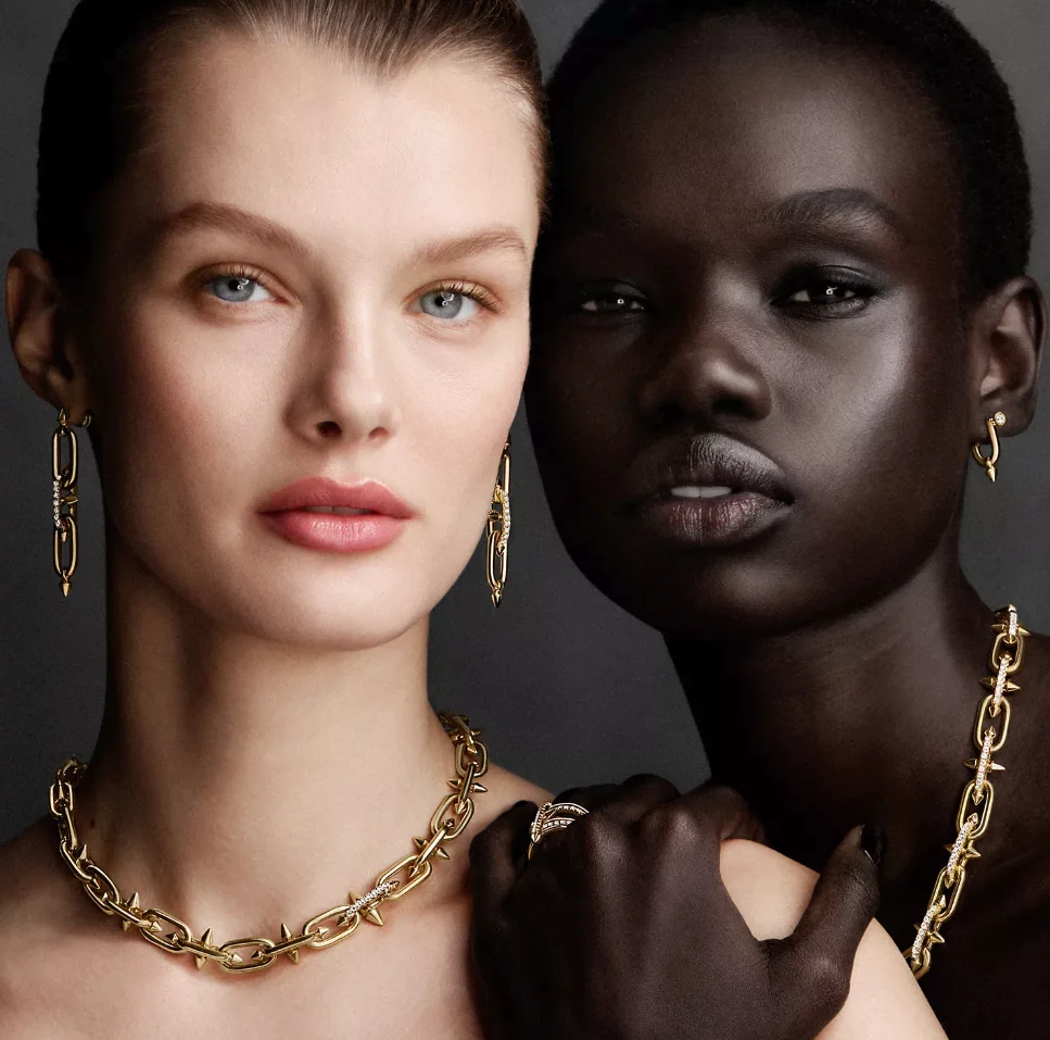 Two models, one light-skinned and one dark-skinned, showcase elegant gold chain jewelry, including earrings, necklaces, and a ring